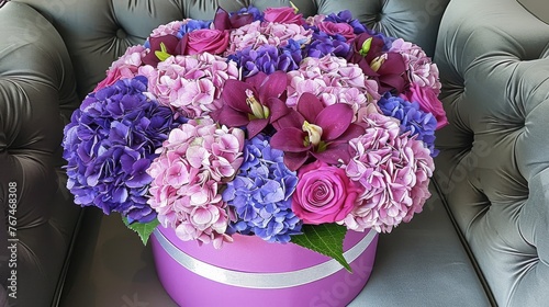  a pink and purple flower arrangement in a purple hat box on a gray leather chair with a silver ribbon around it. photo