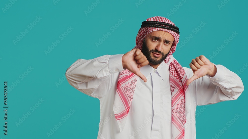 Muslim person gives thumbs down and feeling displeased about something, indicating arabic culture with traditional robe and headscarf. Young adult showing refusal and negative gesture.