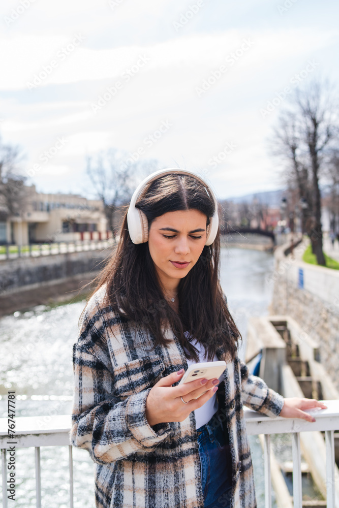 One young girl is listening to music on her wireless headphones and using her phone outdoors on a sunny day
