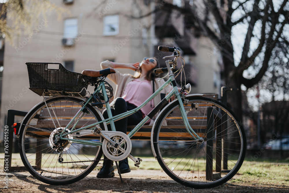 A relaxed young woman taking a break on a wooden bench with a classic bicycle in an urban park.