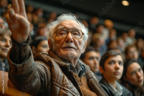 An elderly man is making a gesture with his hand in the air, sitting among a crowd of people at an event. His wrinkled thumb is raised, entertaining the audience of fans