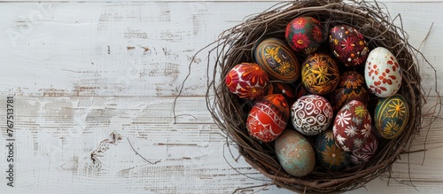 Easter eggs adorned with wax-resist dyeing method, known as Pysanky, displayed in a nest on a white wooden surface. The style is a traditional craft popular in Eastern European regions, photo