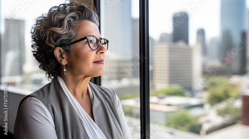 mature optimistic latina hispanic businesswoman woman female executive CEO in corporate modern office thinking contemplating and looking out window skyscraper cityscape daytime with