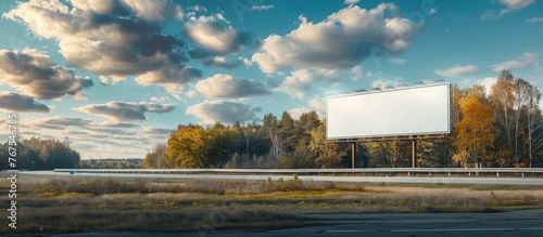 A large vacant mock-up billboard is seen alongside a highway, set against a backdrop of a forest, blue sky, and picturesque clouds.