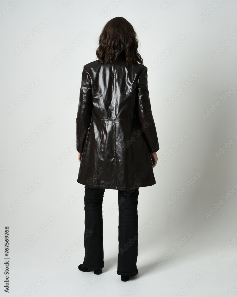 Full length portrait of brunette woman wearing long leather trench coat and black boots. Standing pose walking away from camera, facing backwards. Isolated silhouette on white studio background.