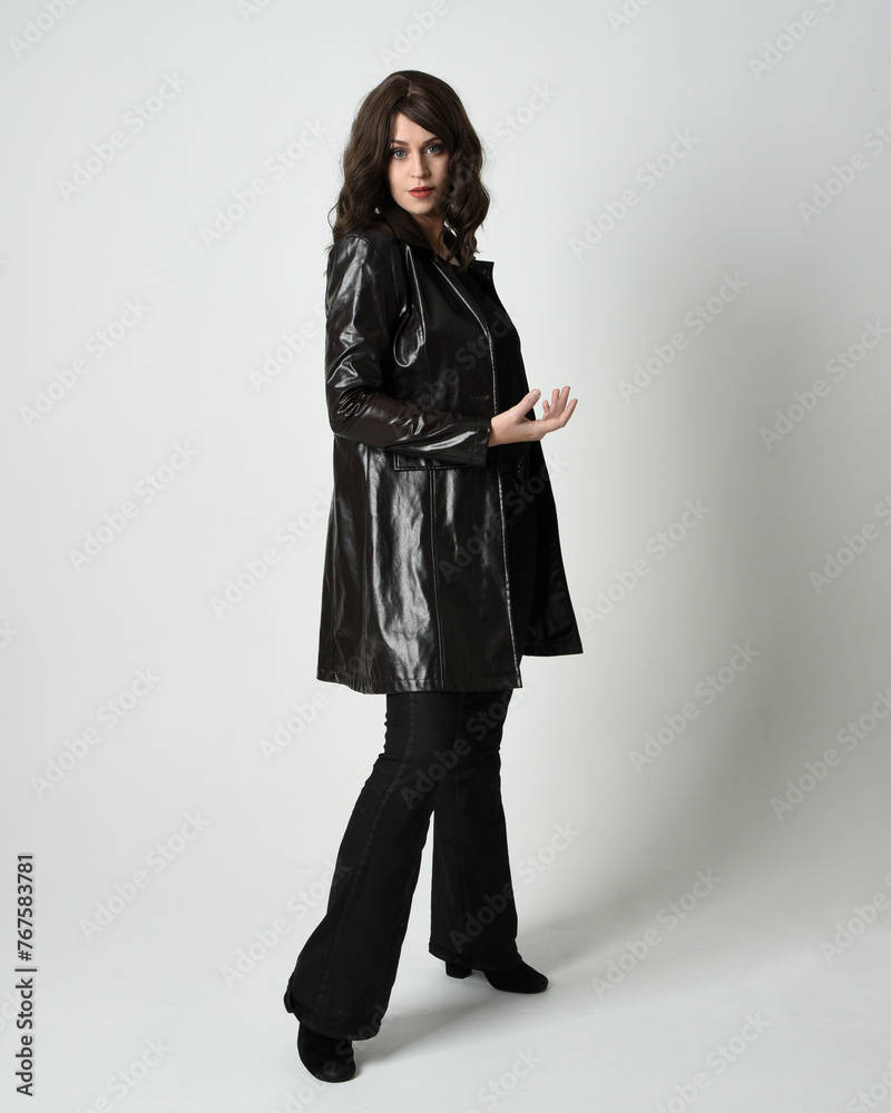 Full length portrait of brunette woman wearing long leather trench coat. Standing pose in side profile, looking back over shoulder, reaching out. Isolated on white studio background. Full length portrait of brunette woman wearing long leather trench coat. Standing pose in side profile, looking back over shoulder, reaching out. Isolated on white studio background.
