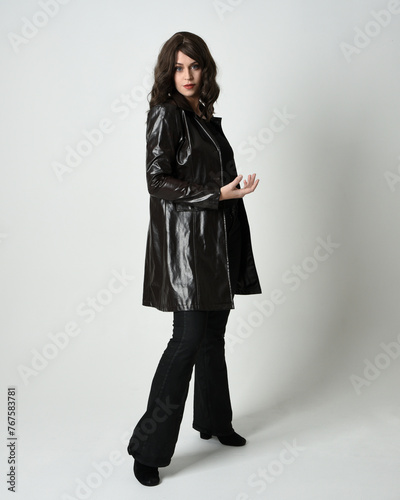 Full length portrait of brunette woman wearing long leather trench coat. Standing pose in side profile, looking back over shoulder, reaching out. Isolated on white studio background.