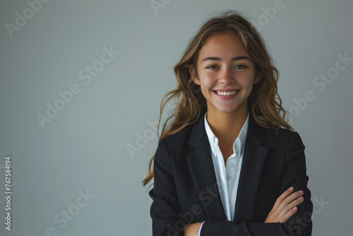 A young woman in a black suit and white shirt is smiling for the camera. She has her arms crossed and is standing in front of a white wall