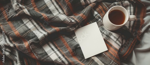 Card and coffee cup placed on a bed with a cozy plaid, offering space for text. Flat lay perspective from above.