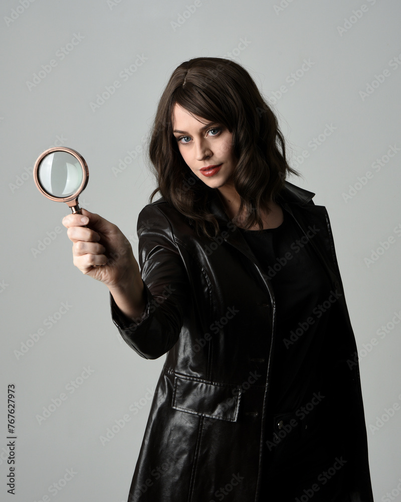 close up portrait of beautiful brunette woman wearing black leather trench coat. Holding detective magnifying glass, reaching out towards camera, searching for discovery. Isolated studio background close up portrait of beautiful brunette woman wearing black leather trench coat. Holding detective magnifying glass, reaching out towards camera, searching for discovery. Isolated studio background