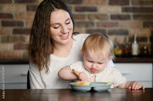  Funny child learns to eat on his own, sitting in his mother's arms in kitchen. Baby independently eats healthy food from plate wit spoon. Concept of self-nutrition, healthy eating, diet. © GRON777