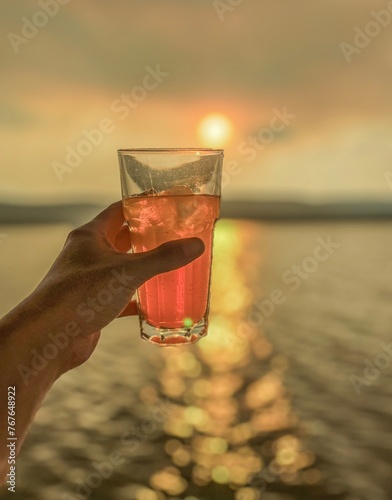 close up of a hand toasting with an orange coloured long drink to a sunset over water