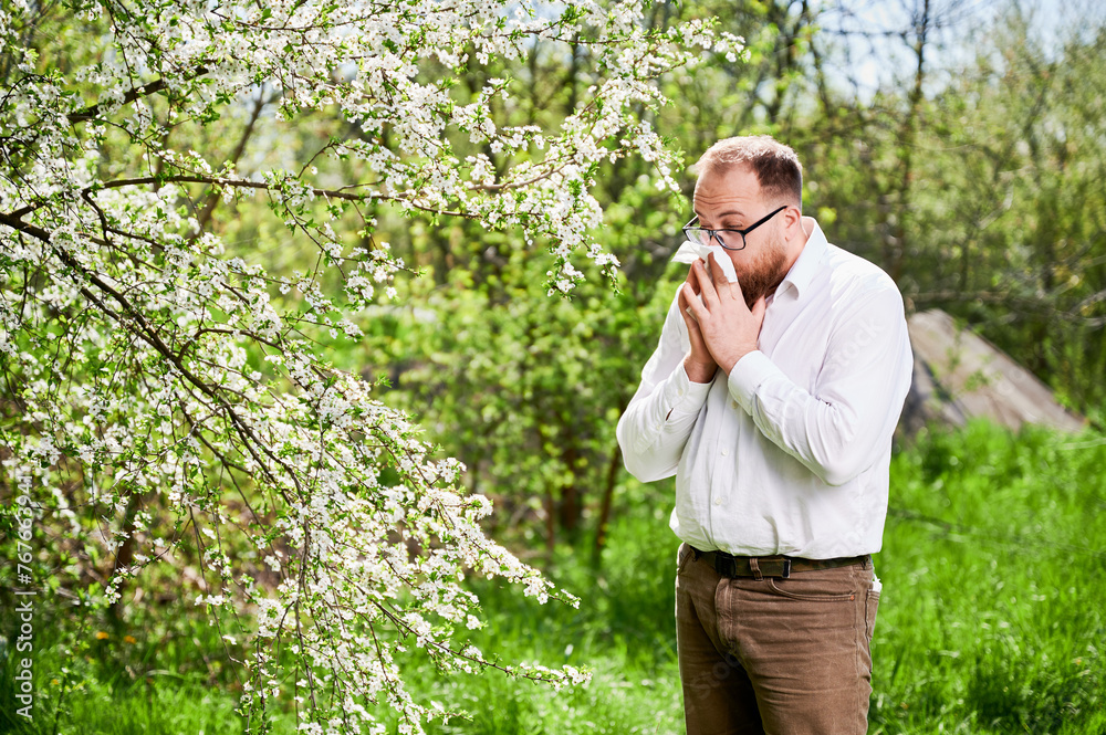 Man allergic suffering from seasonal allergy at spring in blossoming garden at springtime. Young man sneezing and blowing nose with nasal handkerchief in front of blooming tree. Spring allergy concept Man allergic suffering from seasonal allergy at spring in blossoming garden at springtime. Young man sneezing and blowing nose with nasal handkerchief in front of blooming tree. Spring allergy concept