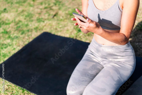 unrecognizable young woman in sportswear kneeling on her mat using mobile phone for a yoga session on the park grass, technology and healthy lifestyle concept © Raul Mellado