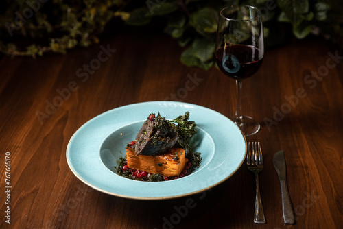 cooked meat with potato lasagna served in blue dish in restaurant with glass of wine on dark background