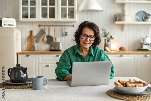 elderly woman in eyeglasses uses laptop sitting in kitchen photo