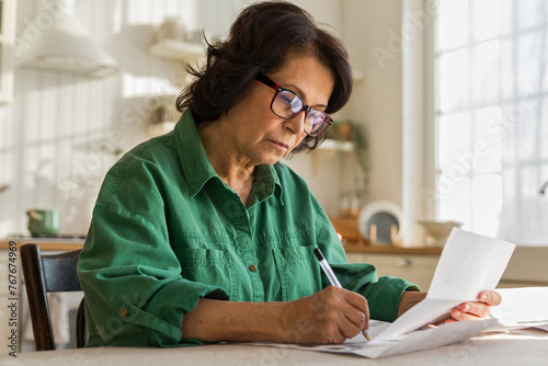 serious elderly woman works with papers and bills, sitting in kitchen photo
