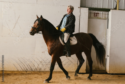 Older man trots on chestnut stallion horse in indoor arena photo