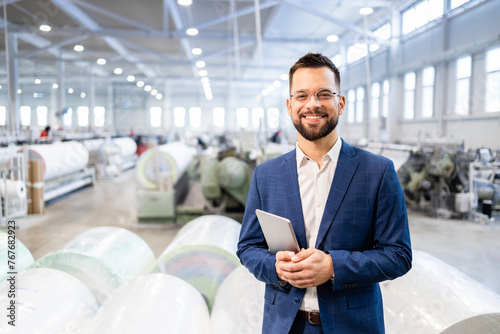 Fotografie Caucasian factory manager or CEO holding tablet computer and checking production results in industrial plant while employees working in background