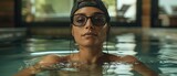 A woman is swimming in a pool wearing a black hat and glasses. She is wearing a black shirt and is in the water
