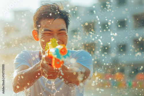 Happy traveler asian man wearing summer shirt holding colorful squirt water gun over blur city, Water festival holiday concept © grapestock