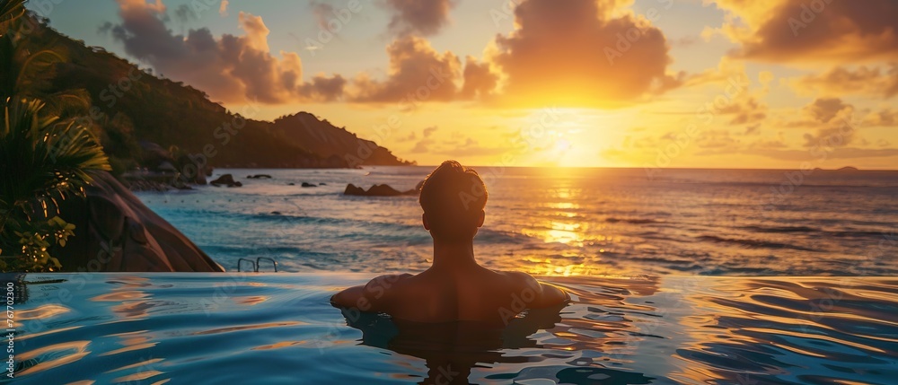 A man is sitting in a pool of water, looking out at the ocean. The sun is setting in the background, creating a warm and peaceful atmosphere