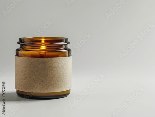 A candle in a jar with a brown band around it. The candle is lit and the jar is sitting on a white surface