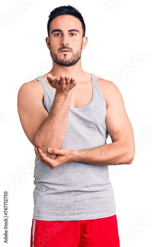 Young handsome man wearing swimwear and sleeveless t-shirt looking at the camera blowing a kiss with hand on air being lovely and sexy. love expression.