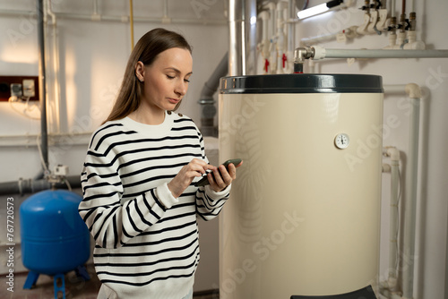 A young woman examines a non-operational boiler with concern, holding a phone in her hand, likely seeking help or planning to call for repair services. The basement setting indicates a residential