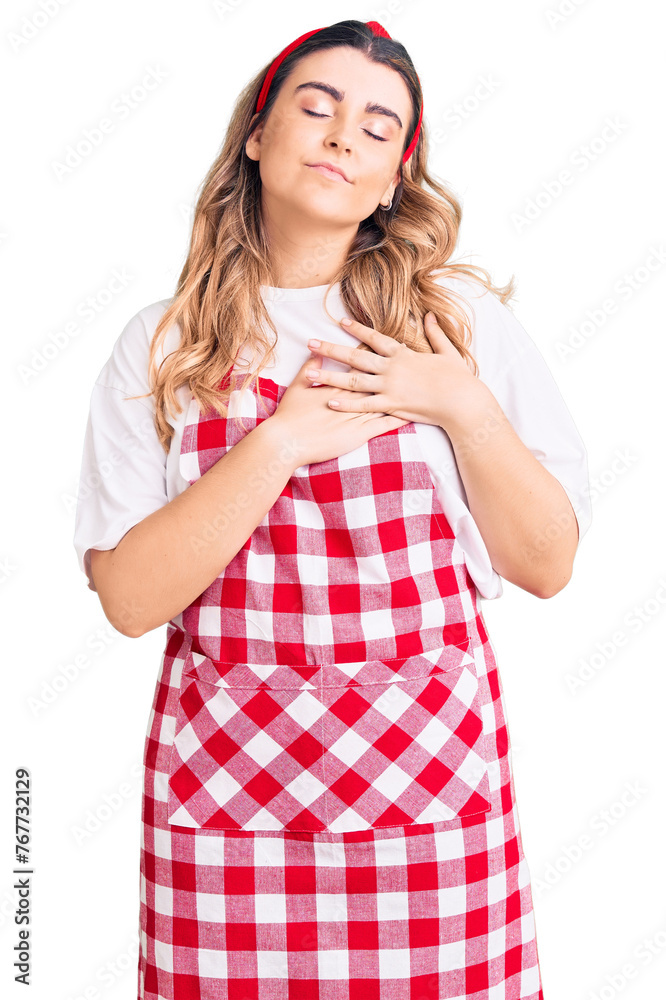 Young caucasian woman wearing apron smiling with hands on chest with closed eyes and grateful gesture on face. health concept. Young caucasian woman wearing apron smiling with hands on chest with closed eyes and grateful gesture on face. health concept.