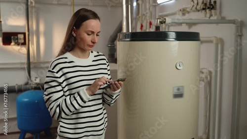 A young woman examines a non-operational boiler with concern, holding a phone in her hand, likely seeking help or planning to call for repair services. The basement setting indicates a residential