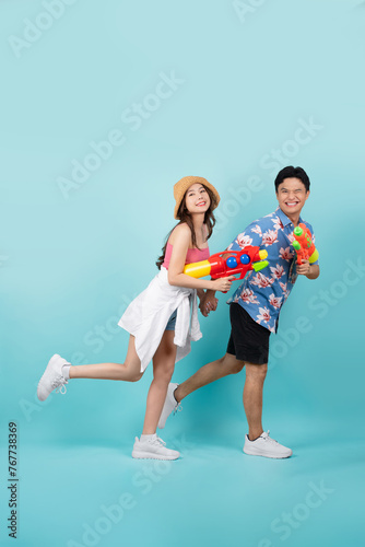 Side view of full length portrait of Young Asian couple walking and holding water gun isolated on blue background