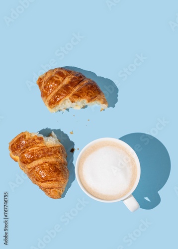 Flat lay of coffee cup and fresh broken croissant on blue background with shadow.