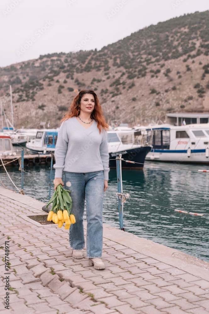 Woman holds yellow tulips in harbor with boats docked in the background., overcast day, yellow sweater, mountains