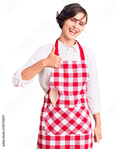 Beautiful young woman with short hair wearing professional cook apron doing happy thumbs up gesture with hand. approving expression looking at the camera showing success.