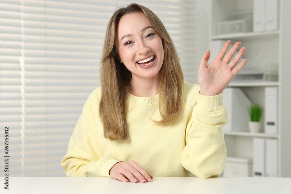 Young woman waving hello during video chat at white table indoors, view from webcam Young woman waving hello during video chat at white table indoors, view from webcam