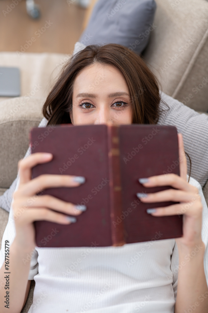 A woman enjoys reading a hardcover book, lying comfortably on a couch in a well-lit living room. A woman enjoys reading a hardcover book, lying comfortably on a couch in a well-lit living room.