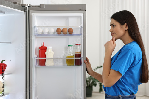 Thoughtful young woman near modern refrigerator in kitchen