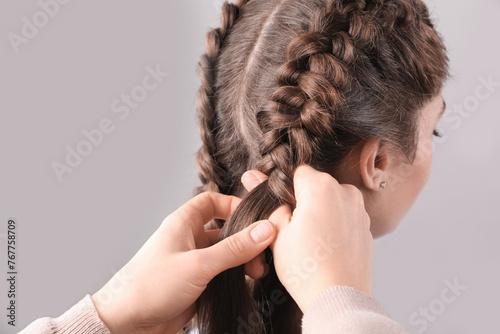 Professional stylist braiding woman's hair on grey background, closeup photo