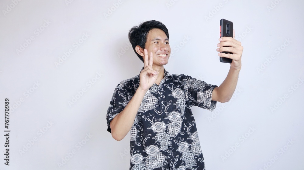 Handsome young Asian man wearing batik is making a video call, taking a selfie using a smartphone Handsome young Asian man wearing batik is making a video call, taking a selfie using a smartphone