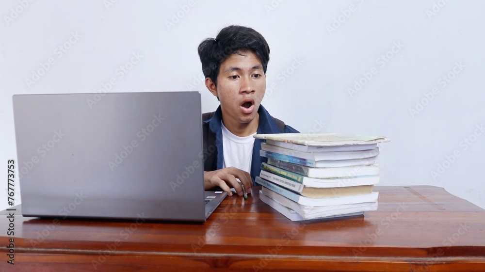 Handsome young male Asian student stressed or shocked studying at a table using a book and laptop Handsome young male Asian student stressed or shocked studying at a table using a book and laptop