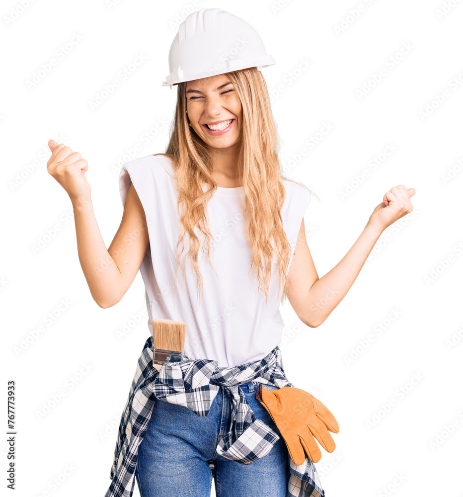 Beautiful caucasian woman with blonde hair wearing hardhat and painter clothes very happy and excited doing winner gesture with arms raised, smiling and screaming for success. celebration concept. Beautiful caucasian woman with blonde hair wearing hardhat and painter clothes very happy and excited doing winner gesture with arms raised, smiling and screaming for success. celebration concept.