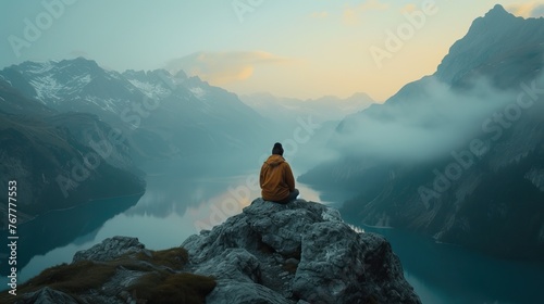 Solitary man in a yellow jacket sitting on a mountain peak overlooking a misty alpine landscape at sunrise. photo
