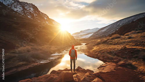 A man stands on a rocky ledge, gazing at the river below as the sun sets. The clouds and sky create a stunning natural landscape, with mountains in the distance and hills leading towards the horizon