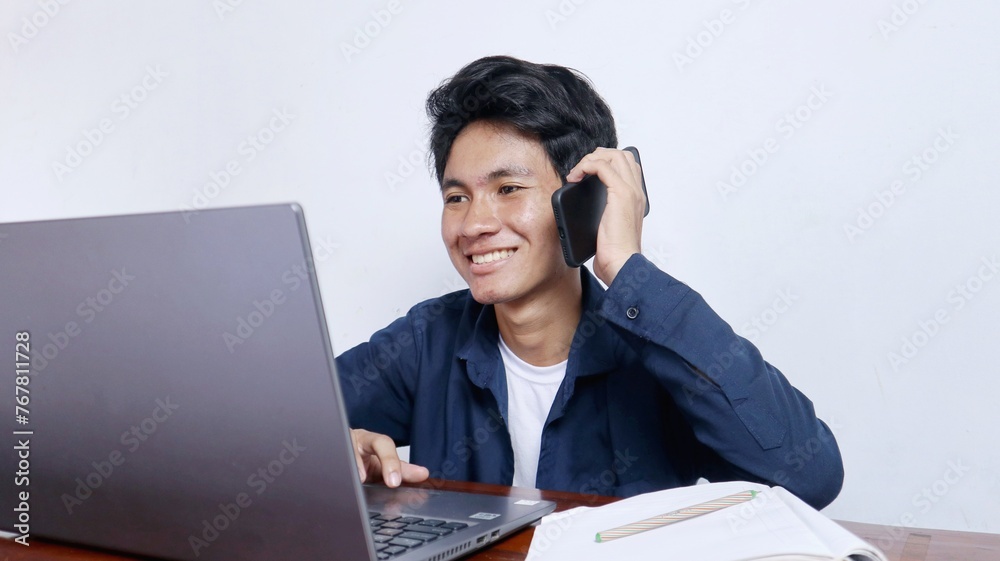 Young Asian male student is studying on a laptop, calling on the phone, doing assignments Young Asian male student is studying on a laptop, calling on the phone, doing assignments