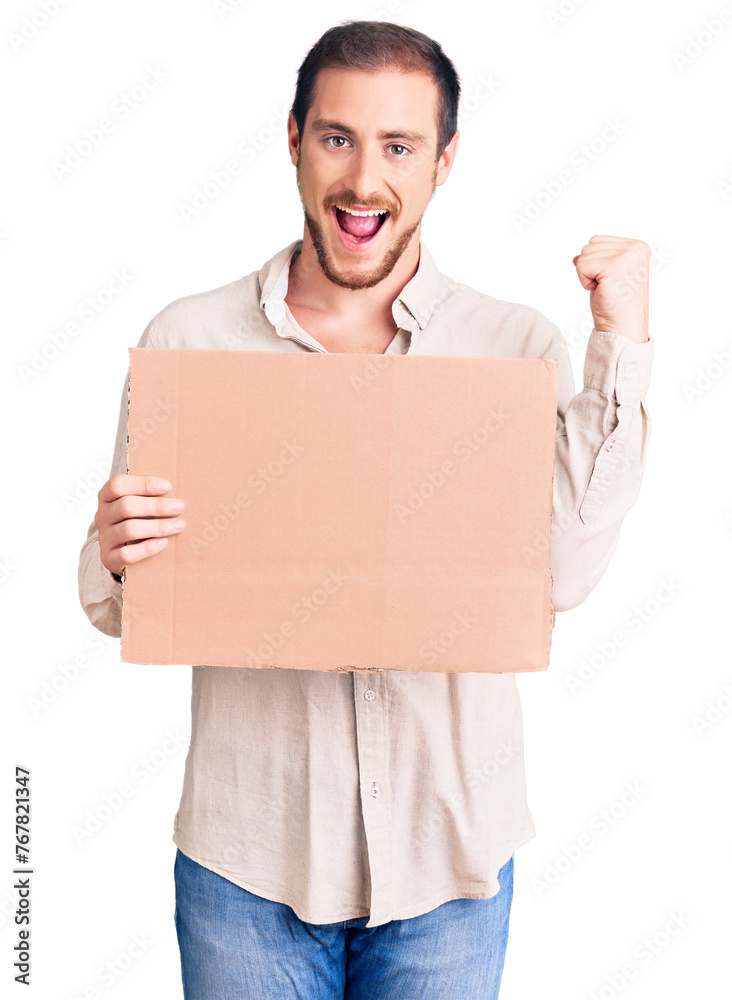 Young handsome caucasian man holding empty cardboard banner screaming proud, celebrating victory and success very excited with raised arms Young handsome caucasian man holding empty cardboard banner screaming proud, celebrating victory and success very excited with raised arms