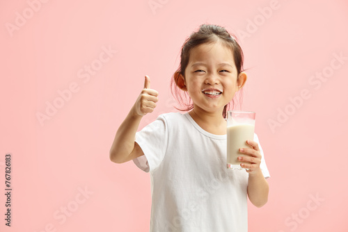 Happy Smiling Little Brunette Child Girl Drinking Milk From Glass With a Trace Frm Milk on Lips, Expresses Pleasure Positively Showing Thumb up Standing On Pink Isolated photo