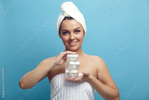Cute smiling young woman in a head towel holding various three moisturizer jars for skin near her face on a blue isolated background. Beauty, spa, skin care.