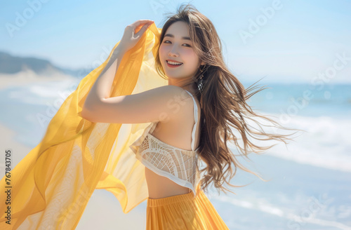 A stunning asian woman with long brown hair, wearing an elegant yellow scarf around her waist and shoulder, stands on the beach under clear blue skies