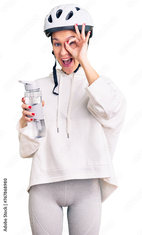 Beautiful brunette young woman wearing bike helmet and holding water bottle smiling happy doing ok sign with hand on eye looking through fingers Beautiful brunette young woman wearing bike helmet and holding water bottle smiling happy doing ok sign with hand on eye looking through fingers